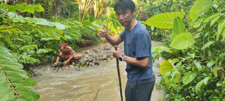 Tour guest giving a thumbs up by the river while a Sikerei prepares medicine in the background in Siberut