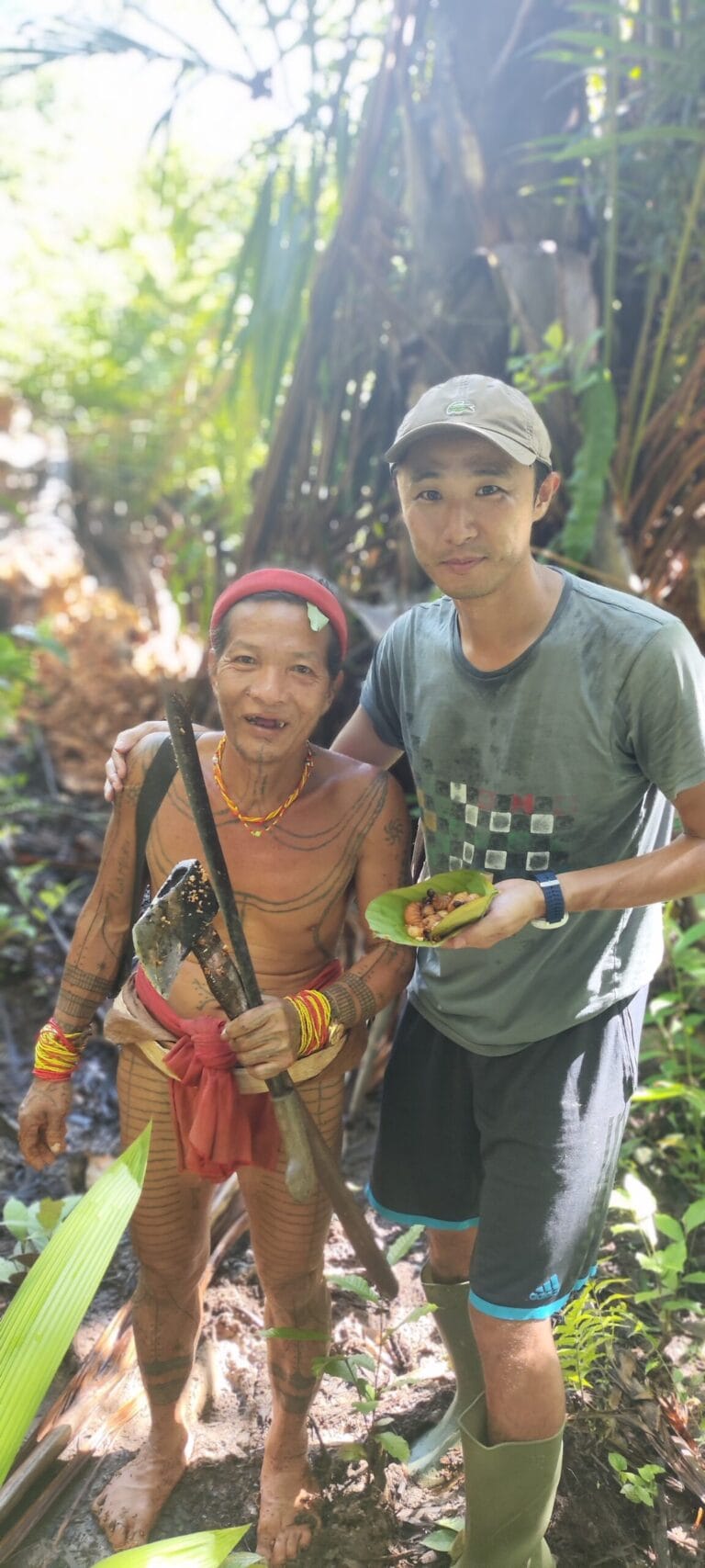 Tour guest with a Mentawai Sikerei holding a forest axe and a cooked forest snack in Siberut