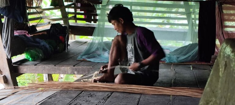 Local Mentawai craftsman weaving rattan strips inside a traditional uma longhouse in Siberut