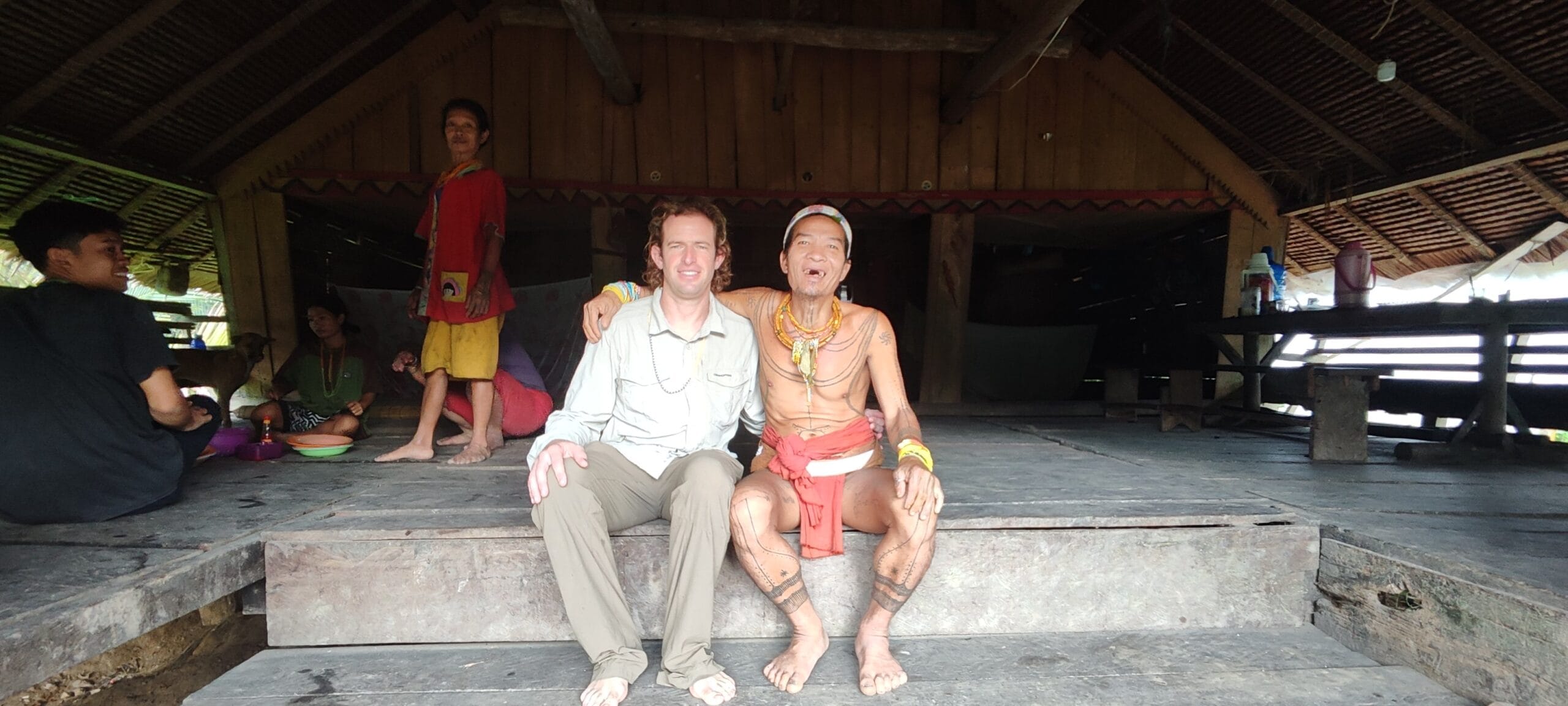 Tour guest sitting with a tattooed Mentawai Sikerei family inside a Siberut uma longhouse