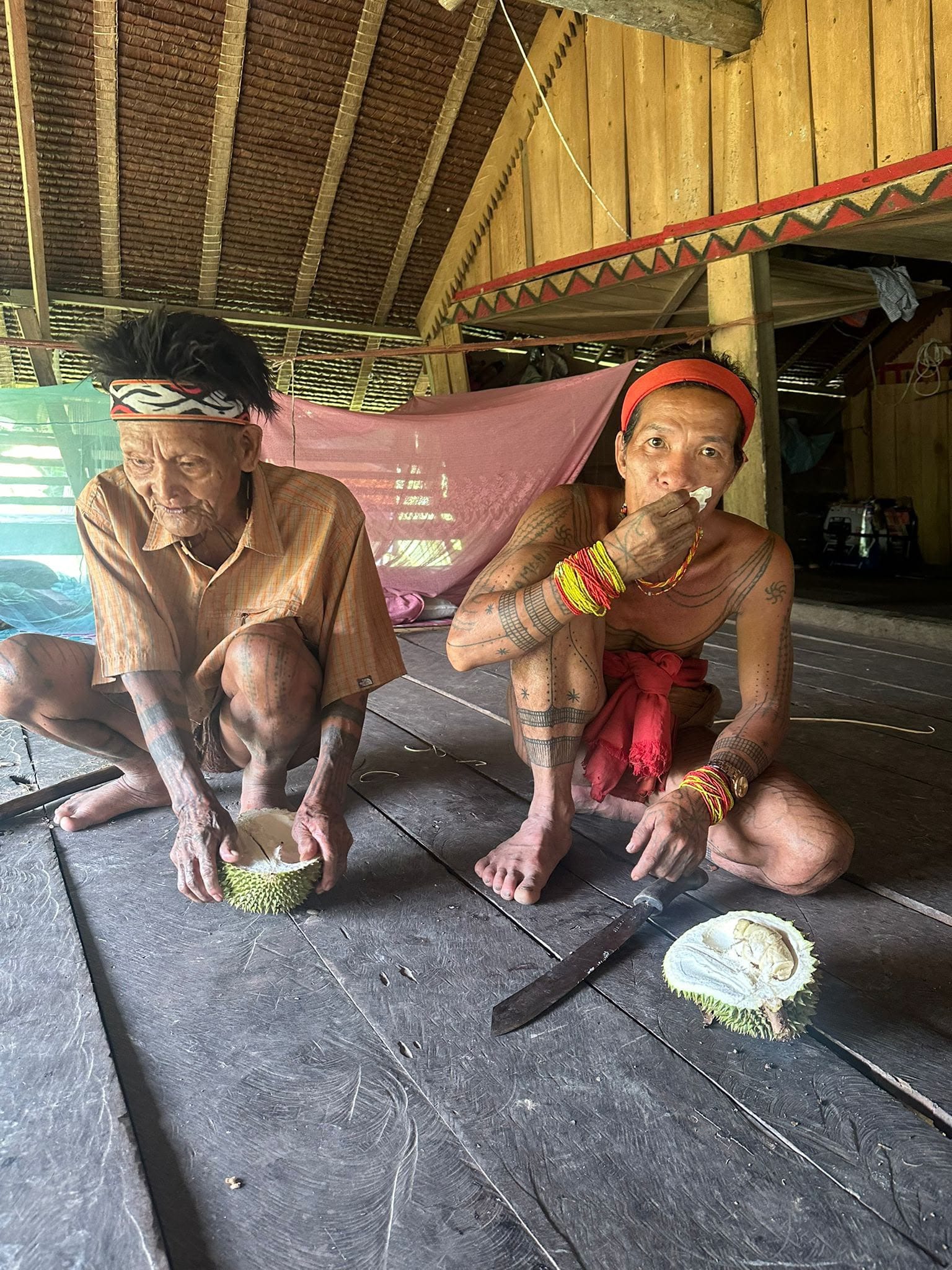 Two elderly Sikerei eating durian, tattoos visible, inside Uma